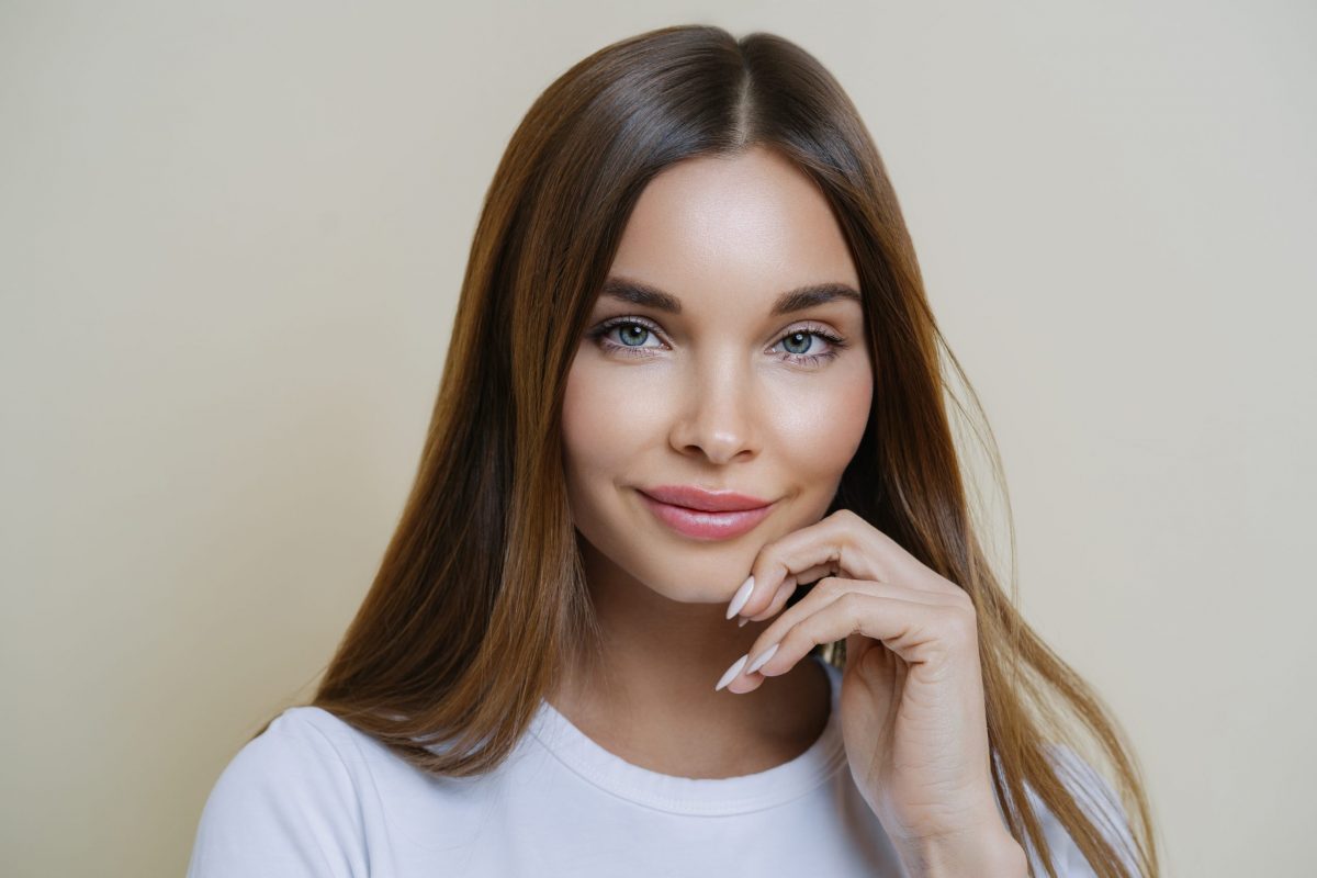 Charming good looking millennial woman keeps hand near face, has healthy glowing skin, dark hair, blue eyes, dressed in casual wear, isolated over brown background. Human face expressions concept