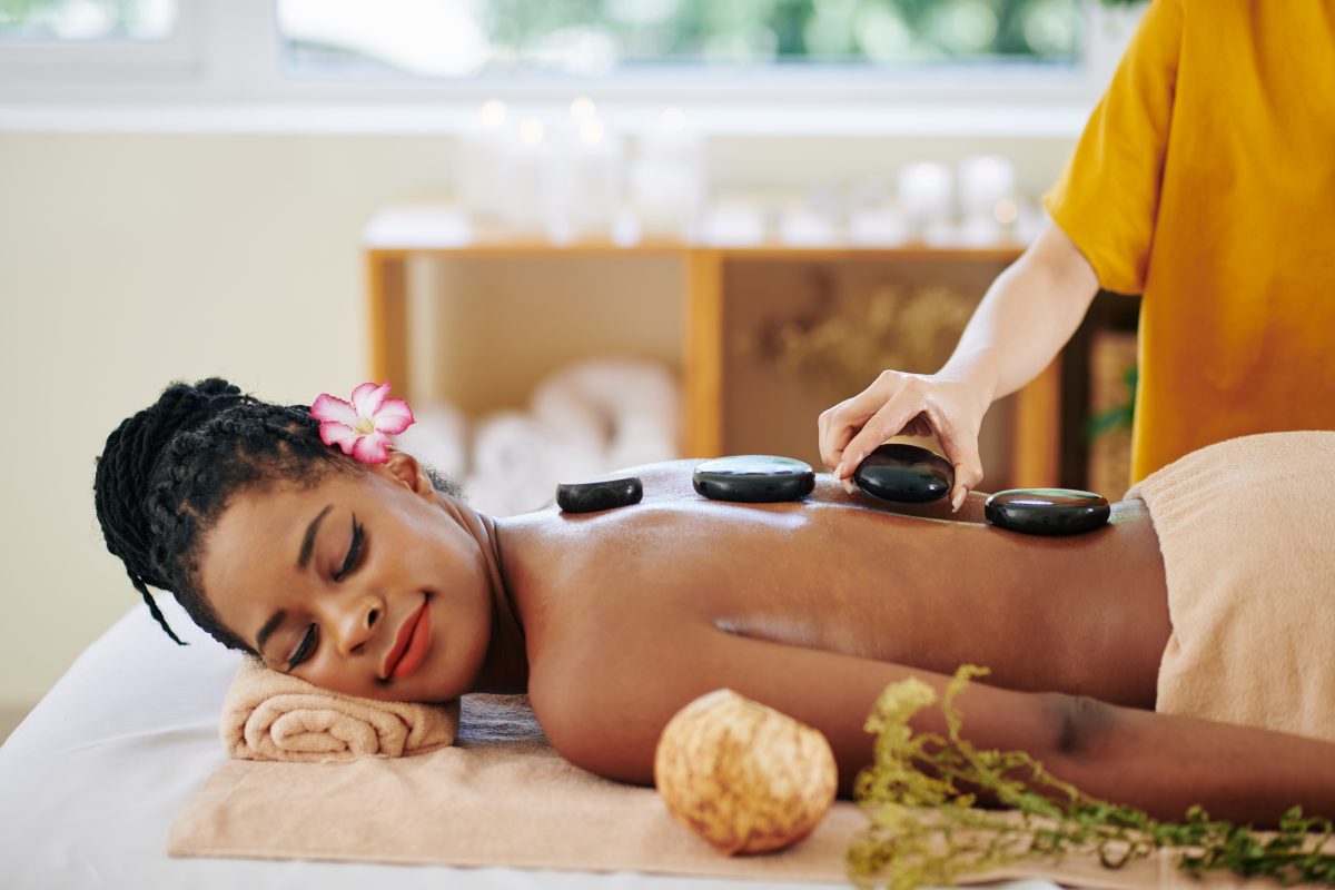 Beautician putting hot basalt stones along the spine of relaxed pretty young Black woman